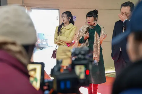 BBC/Hosu Lee Three actors in costume on set pull elaborate facial expressions as they are filmed by a camera.