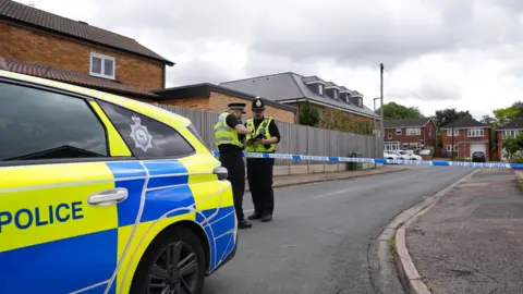 Dawid Wojtowicz/BBC The side of a police car in front of two police officers conferring over police tape cordoning off a road of new build and red brick houses