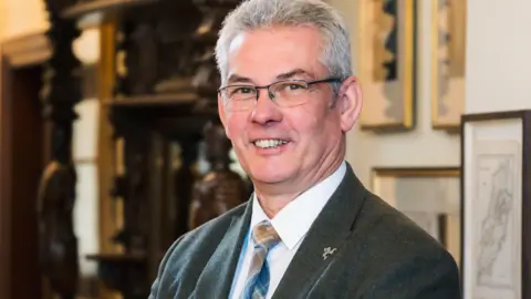 IOM GOVERNMENT Tim Crookall, who has grey hair and is wearing glasses and grey suit with white shirt and Manx tartan tie.