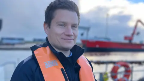 Fiona Irving / BBC A white man with short, dark hair smiles at the camera. He is wearing a blue coat and an orange life jacket. In the middle-ground is some white railings and a life-ring. In the distance is a large red boat.