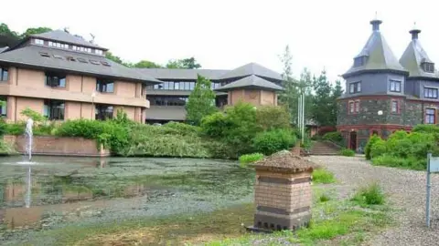 Geograph | Oliver Dixon Powys county hall is a series of two-storey light red brick buildings and a grey brick building with two spires, all behind a pond and an ornamental garden 