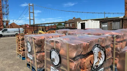 Outdoor view of William Blyth's traditional yard with multiple pallets of terracotta roof tiles wrapped in clear plastic and stacked in the foreground. The packaging displays the William Blyth logo and product labels. Industrial buildings, a white van, and a forklift are visible around the yard. In the background, a large suspension bridge - the Humber Bridge - spans across the scene beneath a bright blue sky with light clouds.