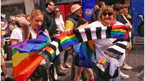 Andi Talbot Two women holding a black and white Newcastle United flag with the rainbow Pride flag interwoven. The woman on the right is siling into the camera and has brown long hair with a full fringe. She is wearing sunglasses and a white t-shirt. The woman on the left is holding a Pride flag and is looking down while she touches her blonde hair.