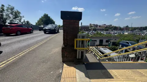 On the left is a road bridge with cars driving over it. On the right, steps lead down to Bedford St John's railway station which will be moved. You can see a train on the track - the front end of it is already passing under the bridge. In the distance is Bedford Hospital and Britannia Road car park, containing hundreds of cars.