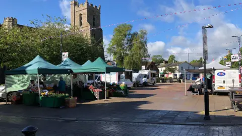Darren Rozier/BBC A market square where various stallholders have set up under green marquee. Vans have been parked by some of the marquees. A church made of red bricks is behind the market. Bunting hangs above the market square. 