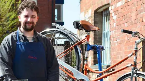 A man in a black jumper and a blue apron stood racing the camera. There is a bike propped up behind him. 