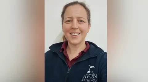 Melanie McPherson Vet Melanie McPherson from Avon Farm vets in Chippenham smiling with he brown hair tied back in a pony tail and wearing a burgundy polo shirt with a navy fleece over the top with a white Avon Farm Vets logo against a white background.