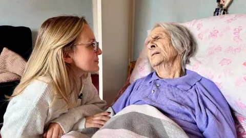 BBC A 98-year-old woman in a purple top is lying in bed with a slightly pained expression on her face.  Her granddaughter is kneeling next to the bed and holding her hand.