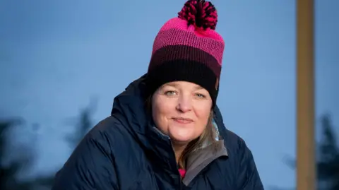 Lianne smiles for the camera in a publicity shot set outside. She is wearing a blue coat and a pink and purple woolly hat.