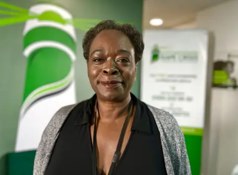 Martin Giles/BBC Dawn Thomas wearing a black V-neck top with a collar and a grey speckled cardigan. She has a black lanyard around her neck. she has short, dark hair, groomed eye brows and a deep brown lip tint on. 