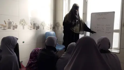 BBC A woman with her face covered teachers girls from whiteboard in a small room 