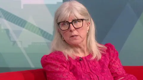 A woman with long blonde hair, glasses and a dark pink top, looks to her right while speaking. She is sitting in a TV studio.