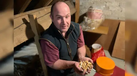 James Christie A man who is partially bald with dark hair is holding the cigarette butts and tips while he sits in an attic. He is wearing a black gilet and burgundy top.