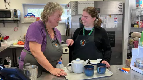 Two women wearing aprons stand at a stainless-steel work surface in a community kitchen with a tea tray, teapot and mugs between them and commercial kitchen equipment in the background.