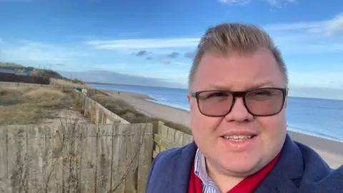 Councillor Scott Dickinson. He has short brown hair and is wearing glasses. He is standing in front of the beach of Druridge Bay.