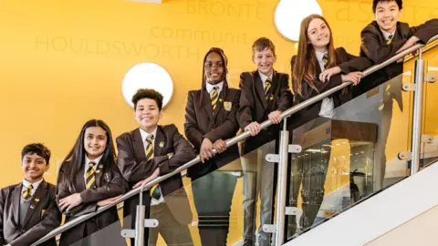 Heckmondwike Grammar School Seven pupils at Heckmondwike Grammar School, in brown blazers and trousers and yellow and brown striped ties, stand on stairs leaning on the handrail. The background wall is yellow. 