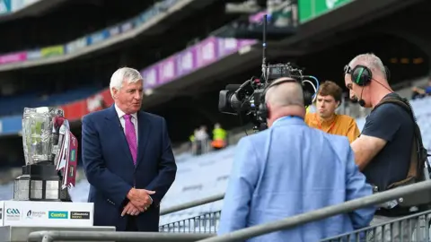 Getty Images A short-grey haired man wears a navy suit, white shirt and pink tie. He speaks into a black TV camera, which is being held by a shirt-grey haired man in a navy t-shirt, wearing a headset. Beside the man in the navy suit is a silver trophy with white and maroon ribbons wrapped around its handle. A bald man in a blue suit and a short-brown haired man in an orange shirt look on from behind the camera. There are rows of empty blue seats behind them all. 
