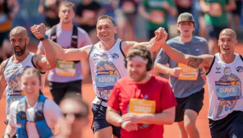 PA Media Three men, Kevin Sinfield in the centre, hold their hands up victoriously. He has short hair and the other men are bald. They are all smiling and wearing matching white running vests with Rob Burrow on them.