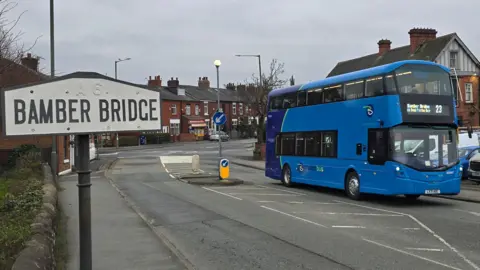 A blue bus driving on a road, with a visable road sign that reads "BAMBER BRIDGE"