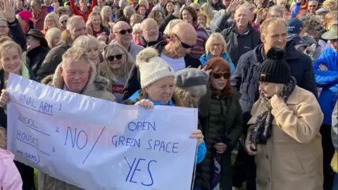 Martin Heath/BBC A group of people standing together, with a couple at the front holding a white banner which reads: Canal Arm 1 School 2 Houses 3 NO / Open Green Space Yes 