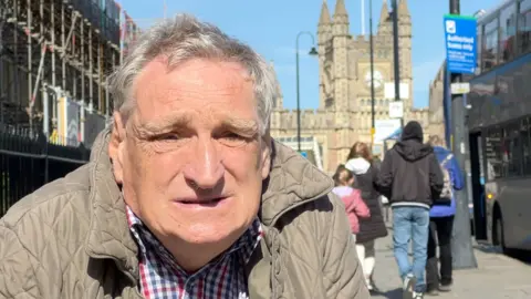 David Redgewell is infant of Bristol Temple Meads. He is wearing a shirt and a beige coloured jacket. Members of the public are walking with their backs to the camera and there is a bus at a stop. 