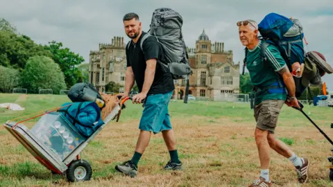 Nathan Roach/Shindig Festival Two men carrying large rucksacks and wheeling other items walk past a stately home