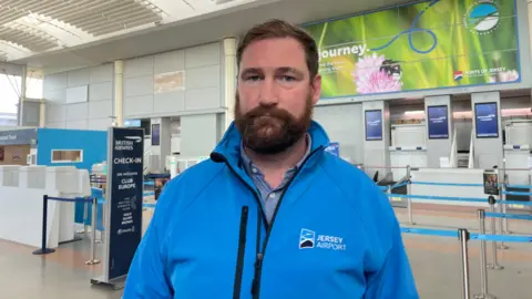 Ashley Maggs - Head of operations Jersey airport wearing a Jersey Airport blue coat standing in front of the British Airways check in area inside the airport.
