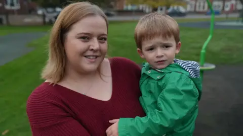A woman holds a young boy in her arms. They are standing in a play park. She looks at the camera and he looks slightly into the distance. 