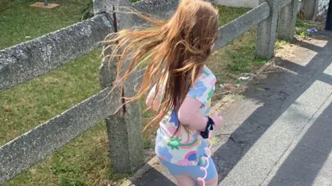 Family handout Nyla running along a paved path on a sunny day. She has long, light brown hair that is flowing behind her as she moves. She is wearing a colourful outfit with a pastel pattern and sandals. A long pink curly leash or cord is attached around her wrist and trails behind her. To her left is a grassy area bordered by a row of weathered concrete posts connected by horizontal rails, and behind that is a tall wooden fence.