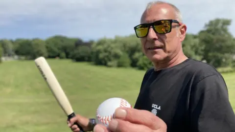 A man wearing a black t-shirt and black sunglasses is holding a rounders bat and ball as he stand in a green park field, with green trees and a blue sky in the background.