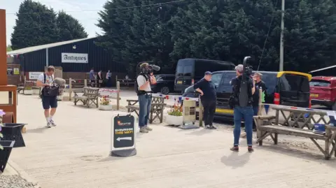Mat Capper A man in a dark top, wearing a flat cap is being filmed by a number of people. He is standing beside a van. There is a large barn behind him with a sign saying antiques