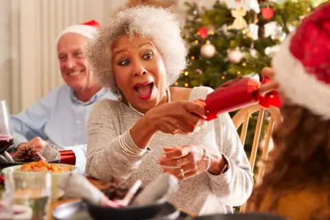 Getty Images A woman with a smile on her faces is pulling a cracker with a family member at the dinner table. She looks as though she is waiting for it to go 'bang'
