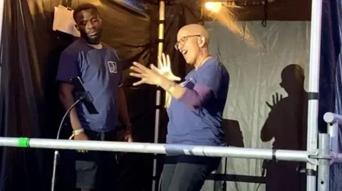 BBC A woman who is doing sign language standing in a gazebo next to the main stage at a festival. She is wearing black jeans and a navy top. A man is also standing to her side.