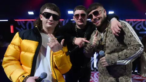 Getty Images The three members of Bad Boy Chiller Crew are wearing dark sunglasses and facing the camera while holding microphones. One is in a bright yellow and black coat on top of a white t-shirt. Another is dressed in black with a gold chain around his neck and wrist. The other is in a patterned beige tracksuit. They are gesturing with their hands and are stood on a stage, lit by neon lights.
