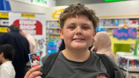 Picture of a boy with brown hair who is smiling and wearing a grey T-shirt and holding a Panini sticker in his hand, in front of the Panini swap shop banner.