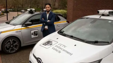 Two cars, one silver and one white, parked outside a building. The text CCTV enforcement vehicle is written along the front bonnet. A man wearing a suit stands in between both vehicles with his arms crossed.