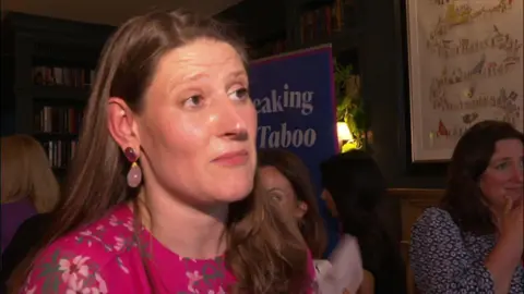 Theo Clarke stands in front of a banner adverting her book. She has shoulder-length dark hair and is wearing a pink dress and has pink earrings.