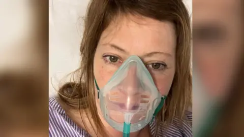 Kerry Fear, wearing a clear mask which helps her breathe. She is dressed in a blue and white striped top and has shoulder length brown hair.