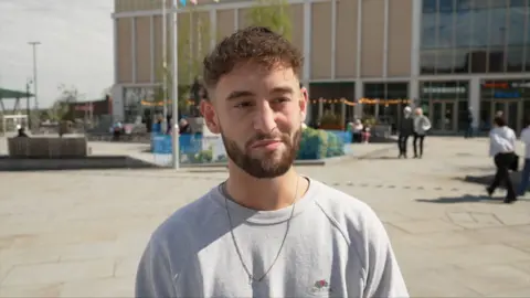 Man with brown hair and beard in a grey top and chain around his neck.