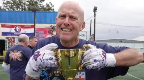 Paul Nicholls Paul Nicholls holding a gold trophy while smiling. He is stood on an astro turf football pitch while wearing white goalkeeper gloves and a blue T-shirt. It is a grey and cloudy day.