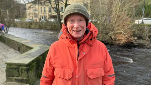 Mark Ansell/BBC A man wearing a red-orange raincoat and green hat. He is standing against the backdrop of a river.