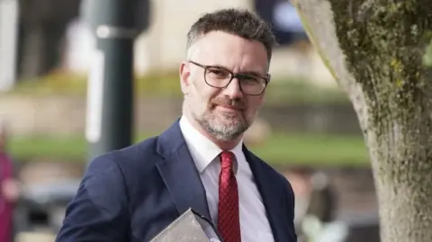 Charles Hanson - he's holding a folder and is wearing a blue suit jacket and a red tie 