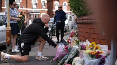 PA Media A man kneels down to read tributes left at the scene. An array of flowers can be seen and more people stand in the background.