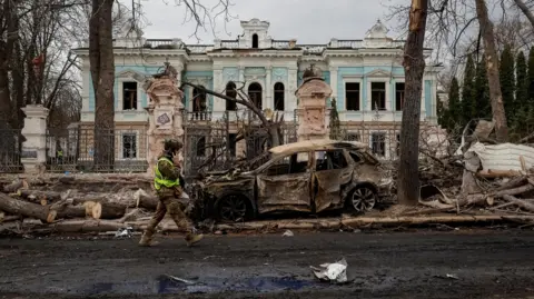 Reuters A Ukrainian serviceman walks at the site of a Russian missile strike in Sumy. A car is wrecked and fallen tree trunks are at the side of the road.