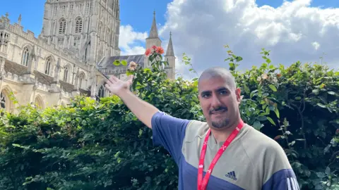 Gardening participant Abteen standing in front of a rose bush in the shadow of Gloucester Cathedral. He is wearing a grey and blue Adidas T-shirt and a red lanyard with white writing on it. He has a shaved head and black facial hair. His arm is pointing upwards to the cathedral.