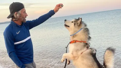 St Thomas More Catholic Voluntary Academy A man with a backwards cap stood next to the sea in a blue top, holding something for his dog, which is on its hind legs looking up.