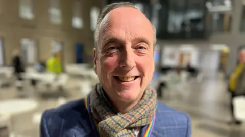 Liberal Democrat leader on Wiltshire Council Ian Thorn smiles at the camera in a blue suit jacket and woolly scarf.
