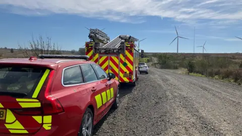 A fire engine parked in front of a fire and rescue car down a rural gravel path. It is day time and wind turbines can be seen in grass fields.