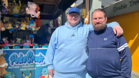 Jack Maclean/BBC Tommy and Stephen Simpson are standing with their arms around each other in front of a market stall. They are both wearing blue tops. There are toys and other merchandise on the shelves of the stall behind them.