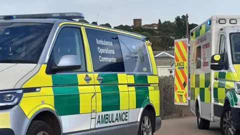 Caroline Robb Two ambulances parked next to each other. The one on the left is more like a van and has "Ambulance Operational Commander" written on the side. The other ambulance on the right has its back doors open. There is a house behind a hedge in the background and a building on top of a hill in the distance.
